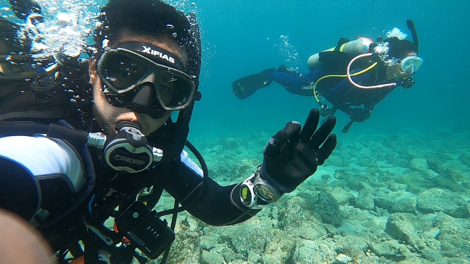 A diver with a regulator in his mouth giving an okay sign, a second diver behind him
