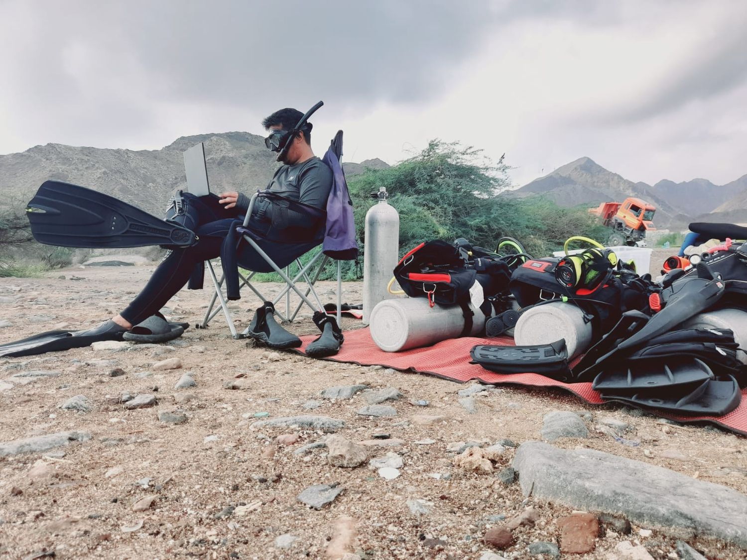 A diver kitted up in a folding chair on the beach between dives, more kit laid out on a mat, mountains behind