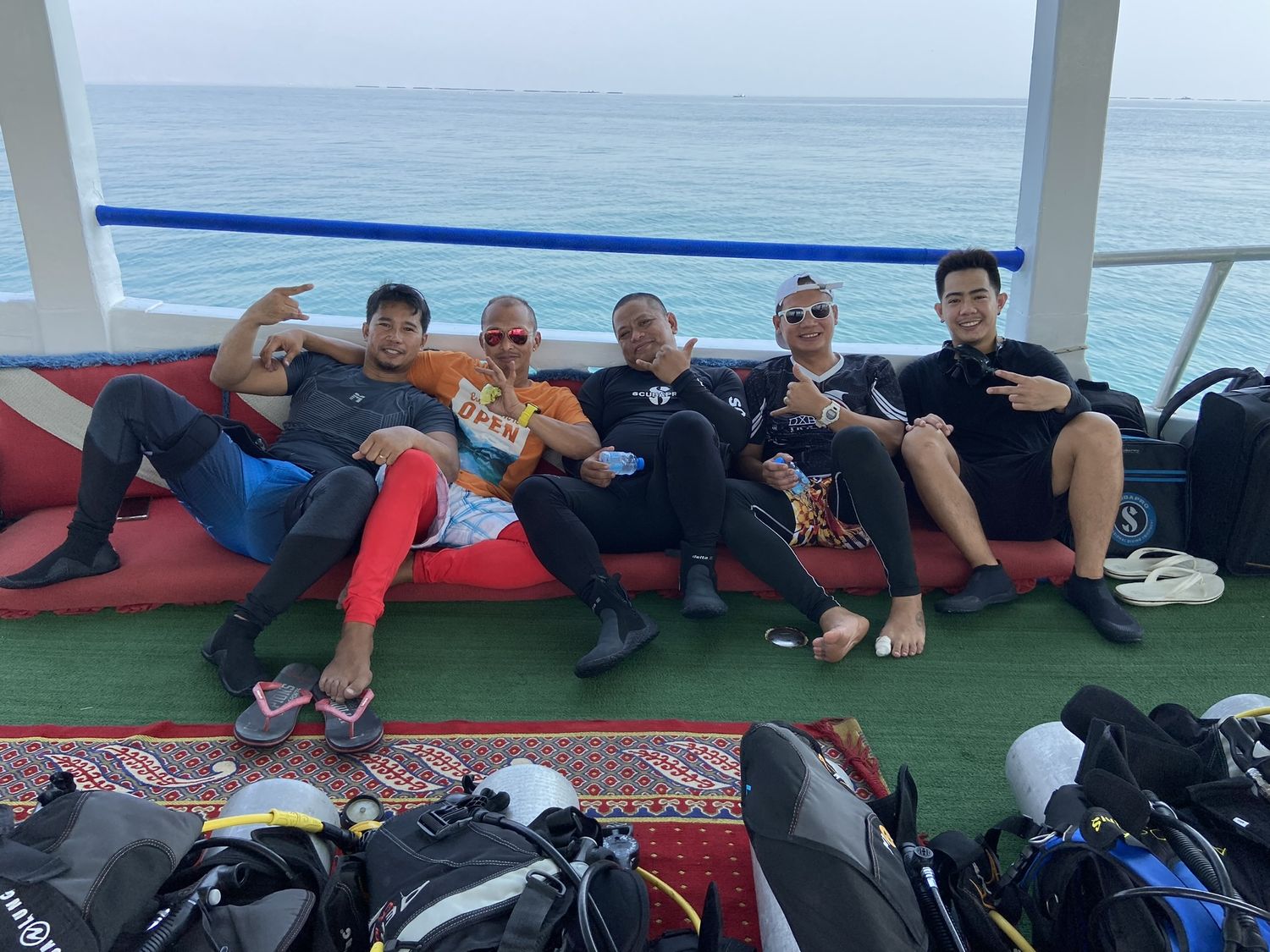 A group of divers sitting along the boat deck in wetsuits, thumbs up, dive bags stowed in front of them
