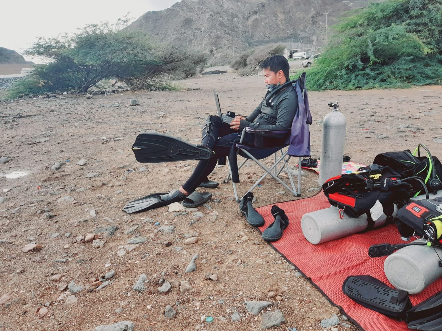 A diver resting in a beach chair between dives with the wetsuit peeled down, kit drying on the mat