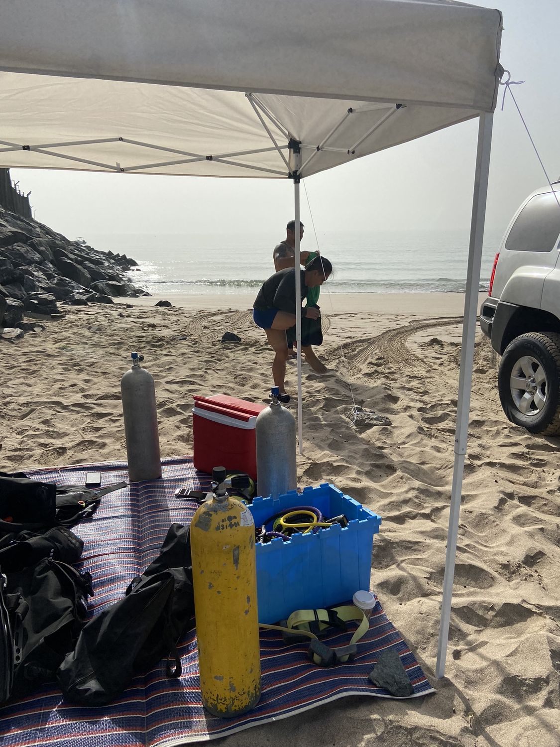 Tanks, a coolbox and dive gear on a mat under a shade canopy on the beach, the sea behind